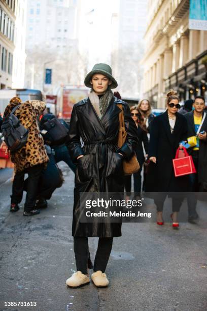Model Cara Taylor wears a green bucket hat, black leather trench coat with pockets, black pants, fuzzy fleece clogs, and a brown corduroy bag after...