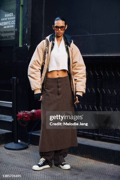 Model Jordan Daniels wears a brown sunglasses, tan puffer jacket, white knit crop top, long brown tweed A-line skirt over brown striped pants, and...