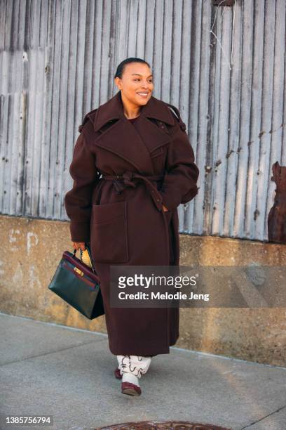 Model Paloma Elsesser wears a long brown belted coat, white print pants, red ballet flats, and carries a dark green and red Hermes Kelly Birkin bag...