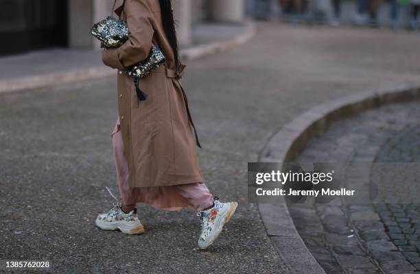 Fashion Week guest seen wearing a brown trench coat, a pale pink long satin dress, a gold glitter handbag and white with flower embroidered sneakers...