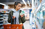 Young woman reading nutrition label while buying diary product in supermarket.
