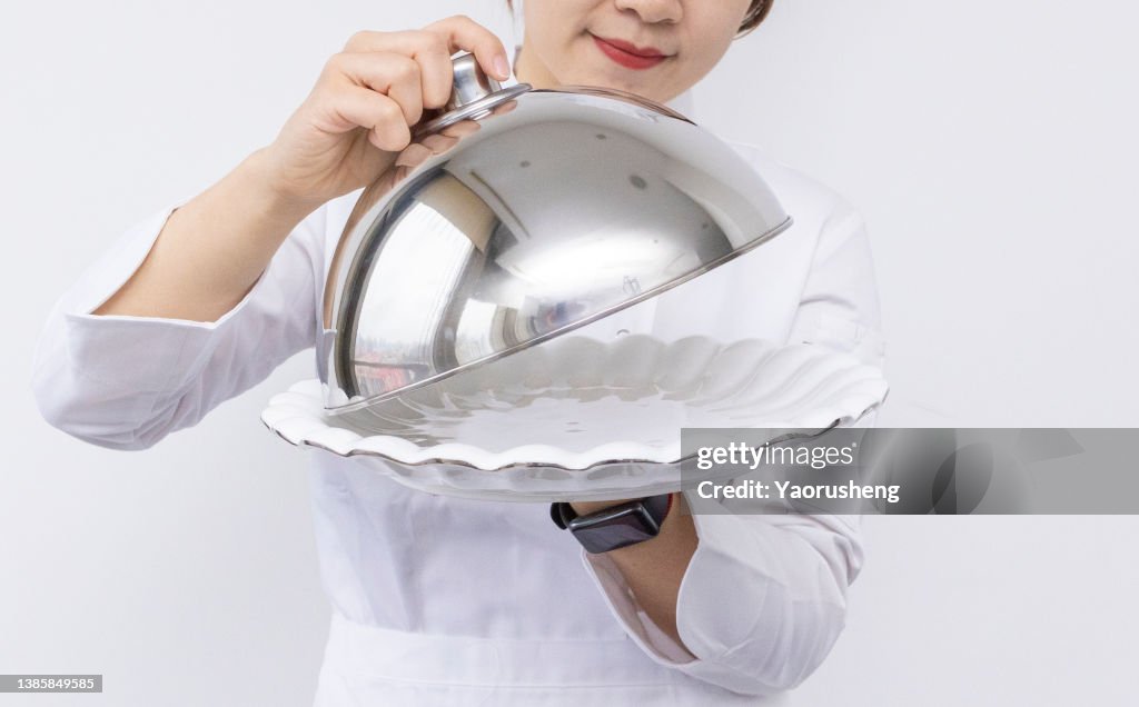 Asian female chef wearing a chef's hat and carrying a domed tray and holding a empty plate. advertisement material
