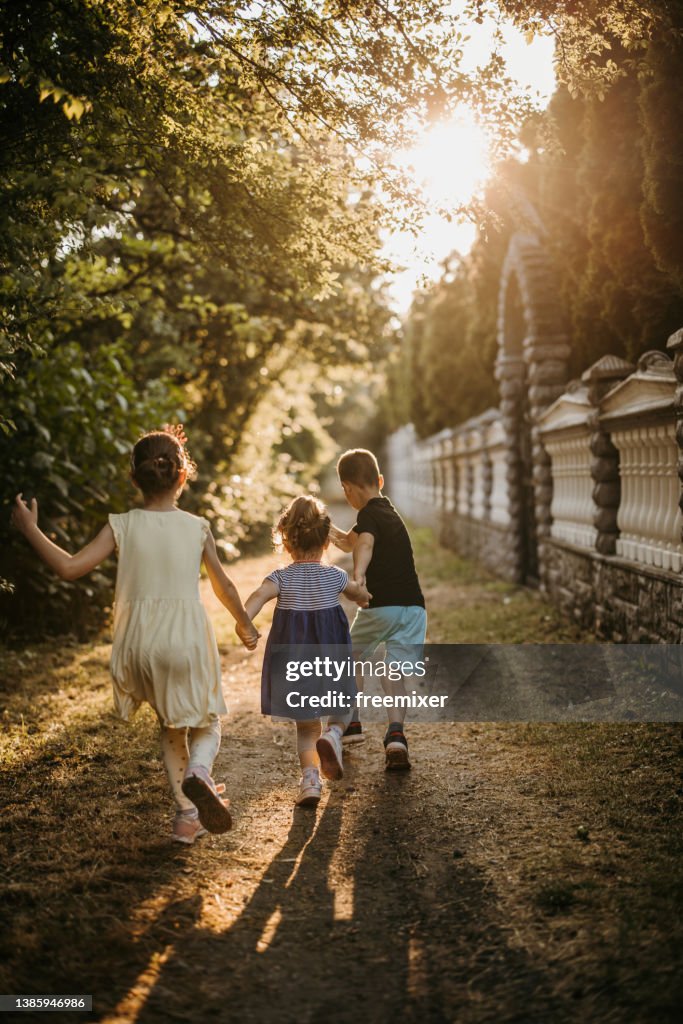 Rückansicht von drei Kindern beim Händchenhalten und Laufen auf dem Fußweg
