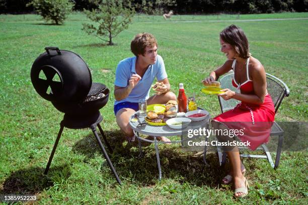 1970s Couple Man And Woman Picnicking In Backyard With Small Charcoal Grill Cooking Hot Dogs And Hamburgers.