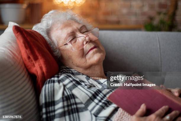 tired senior woman sitting on sofa at home and sleeping when holding book. - sober leven stockfoto's en -beelden