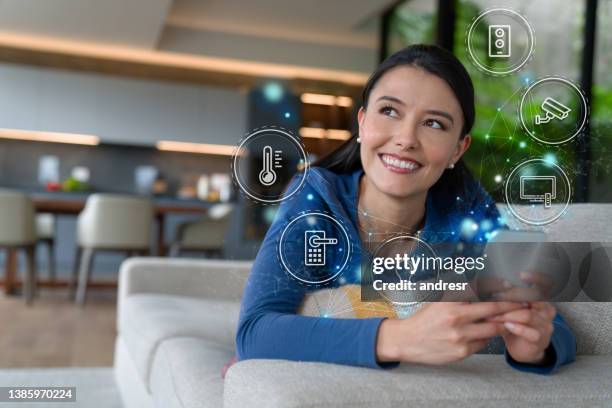 mujer controlando su hogar inteligente desde su teléfono celular mientras está acostada en el sofá - connected home fotografías e imágenes de stock