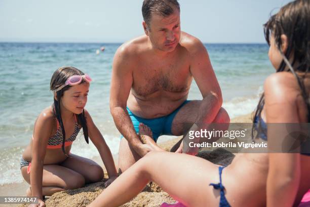 serious man taking care of his daughter's swollen ankle on the beach - tween girl swimsuit stockfoto's en -beelden