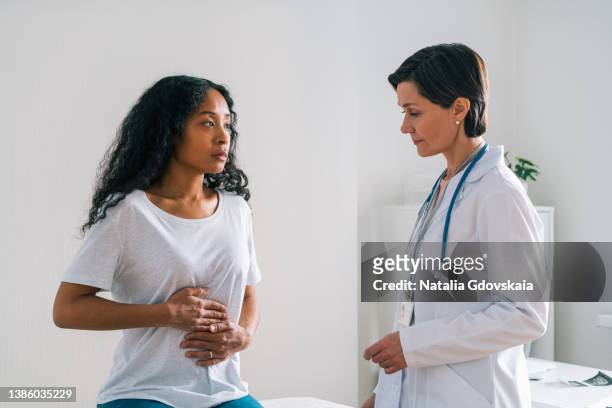 young african-american patient complaining about belly ache to mature female doctor - stomachache fotografías e imágenes de stock