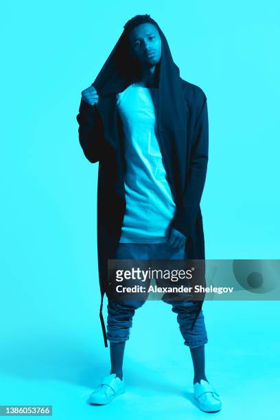 male portrait of black african-american man at the studio in neon lighting. color background, copy space. serious handsome person wear hoodie posing for camera. photo with sneakers and jeans - man and his hoodie imagens e fotografias de stock