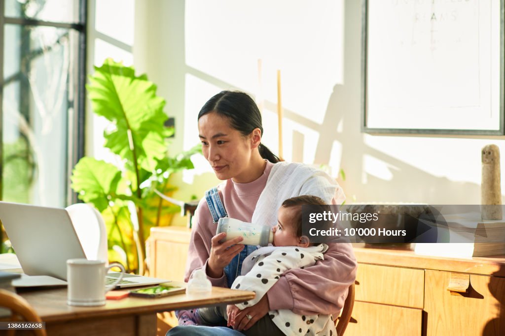 Chinese business woman working at home and bottle feeding baby daughter