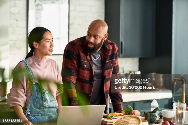 multiracial couple in kitchen preparing dinner with laptop on worktop - woman portrait kitchen laptop stock-fotos und bilder