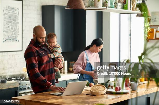 mother preparing meal in kitchen as father hold baby boy and uses laptop - familie laptop stock-fotos und bilder