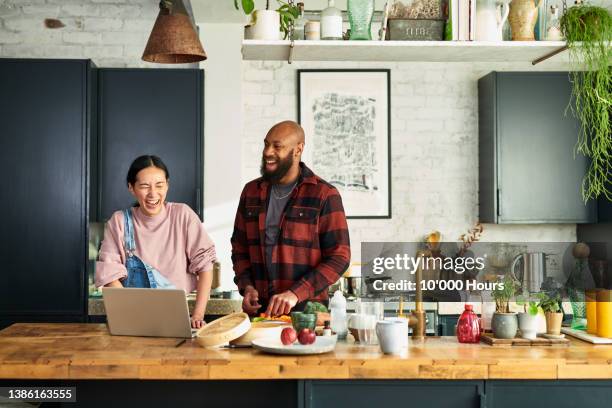 mature chinese woman and black mid adult man preparing food and laughing in kitchen preparing food with laptop on worktop - cooking kitchen stock-fotos und bilder