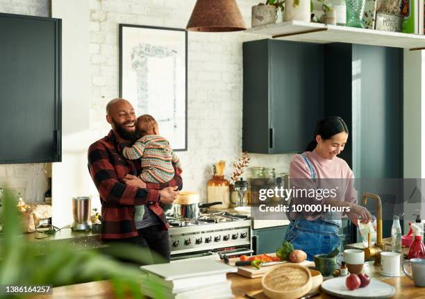 multiracial couple in modern kitchen, black man holding baby boy, mother preparing bottle - domestic chores photos et images de collection
