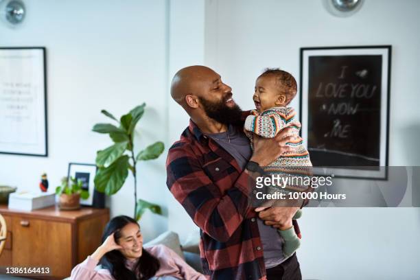 cheerful black father with shaved head and beard holding baby son with mother in background - mann bart portrait mit kind stock-fotos und bilder