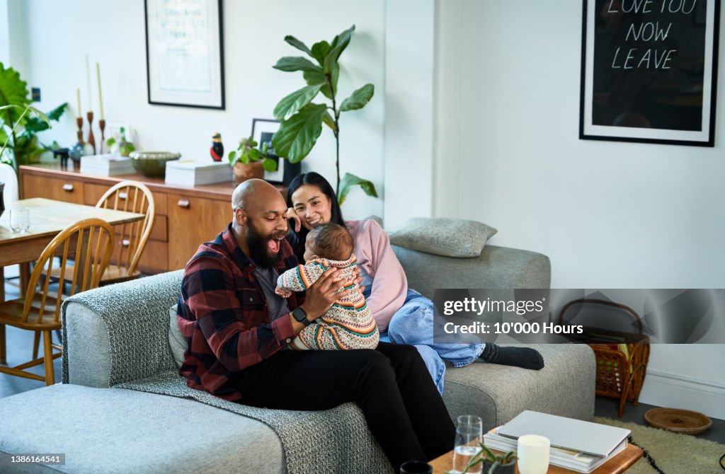 Multiracial parents sitting on sofa with baby son on black mid adult father's lap and Chinese mature mother smiling