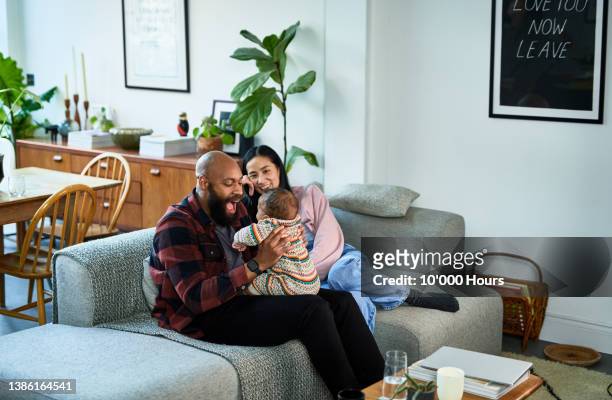 multiracial parents sitting on sofa with baby son on black mid adult father's lap and chinese mature mother smiling - frau anfang 30 stock-fotos und bilder