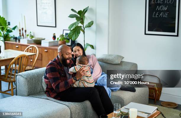 multiracial parents sitting on sofa with baby son on black mid adult father's lap and chinese mature mother smiling - baby at home stock pictures, royalty-free photos & images