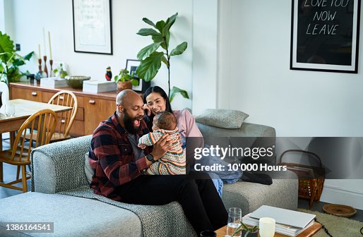 Multiracial parents sitting on sofa with baby son on black mid adult father's lap and Chinese mature mother smiling Multiracial parents sitting on sofa with baby son on black mid adult father's lap and Chinese mature mother smiling