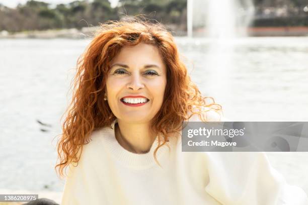 donna dai capelli rossi con un sorriso mentre posa davanti alla telecamera in una giornata di sole. - older woman happy smile foto e immagini stock