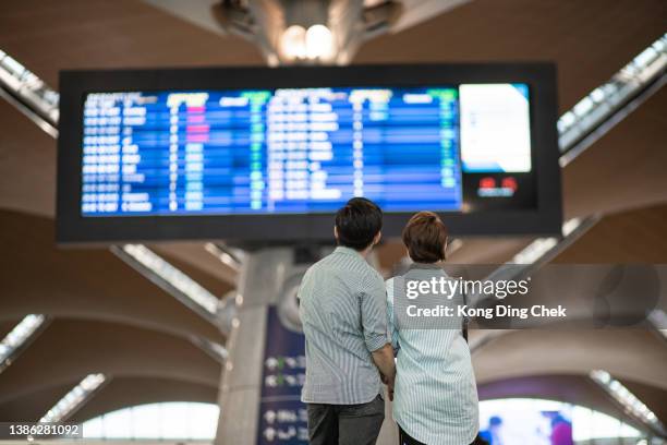 vista trasera. pareja china asiática comprobando la hora de embarque en el aeropuerto.  esperando en frente de la tabla de salida de llegada. - cancelación fotografías e imágenes de stock