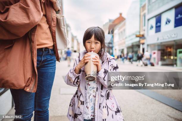 lovely little girl smiling joyfully at the camera & enjoying an iced drink while walking in downtown district with her mom - consumer journey stock pictures, royalty-free photos & images