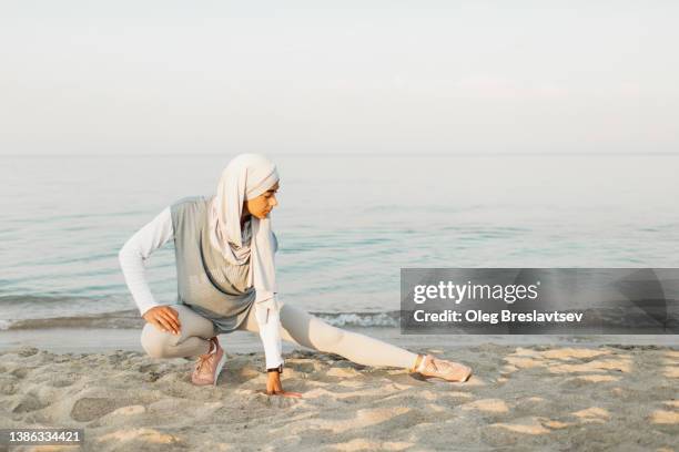 sportive muslim woman in hijab exercising outdoors on beach with sea view, stretching legs with beautiful morning sunlight - muslim woman beach - fotografias e filmes do acervo