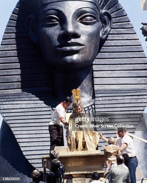 Elizabeth Taylor , British actress, have her costume of ceremonial robes adjusted as she sits before a large Egyptian statue on the set of the film,...