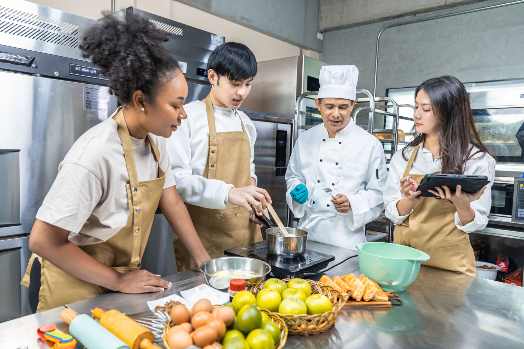 Students In Cookery Class Mixing Ingredients For Recipe In Kitchen.Male And Female young Students With chef Teacher Preparing Ingredients For Dish In Kitchen Cookery Class. Students In Cookery Class Mixing Ingredients For Recipe In Kitchen.Male And Female young Students With chef Teacher Preparing Ingredients For Dish In Kitchen Cookery Class.