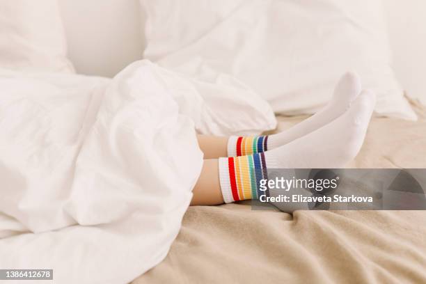 one gay man with white socks on his feet with rainbow symbols of the lgbt community is lying on a bed under a white blanket in a cozy room at home. copy space - feet in bed stock-fotos und bilder