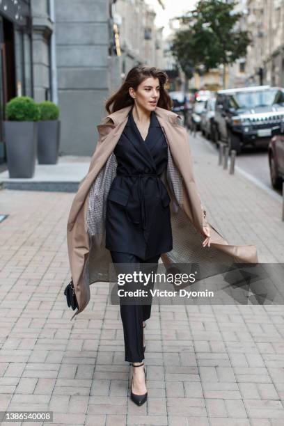 view of a young model in a light brown coat walks on the street and looks away from the camera. - beige coat stock-fotos und bilder