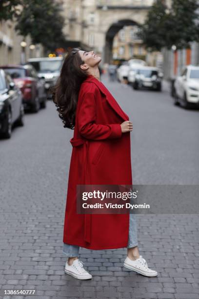 a young brunette stands in a red coat, looks up to the sky and smiles - roter gürtel stock-fotos und bilder