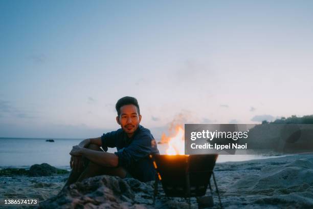 man enjoying campfire on beach at dusk - fuego al aire libre fotografías e imágenes de stock