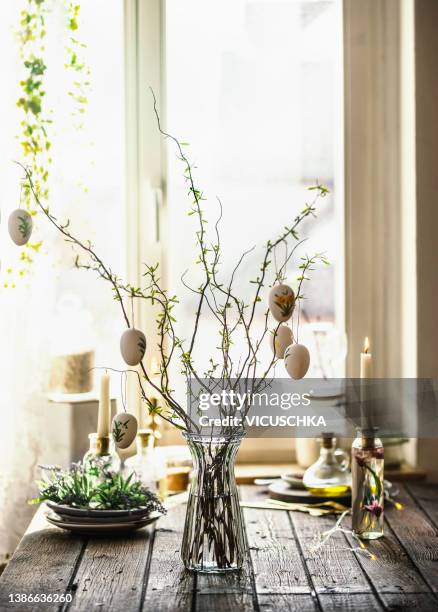 easter branches bunch with hanging easter eggs in glass vase on rustic wooden kitchen table with kitchen utensils, candle and decoration at window background - frühling deko stock-fotos und bilder