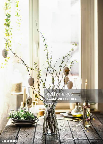easter branches bunch with hanging easter eggs in glass vase on rustic wooden kitchen table with kitchen utensils, candle and decoration at window background - paastafel stockfoto's en -beelden