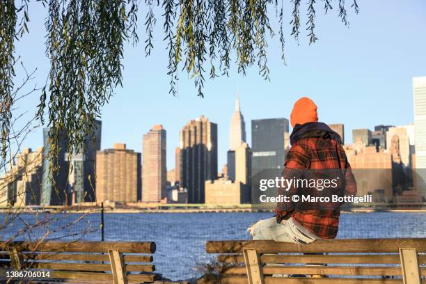 rear view of man sitting on a bench looking at new york city - long island city stockfoto's en -beelden