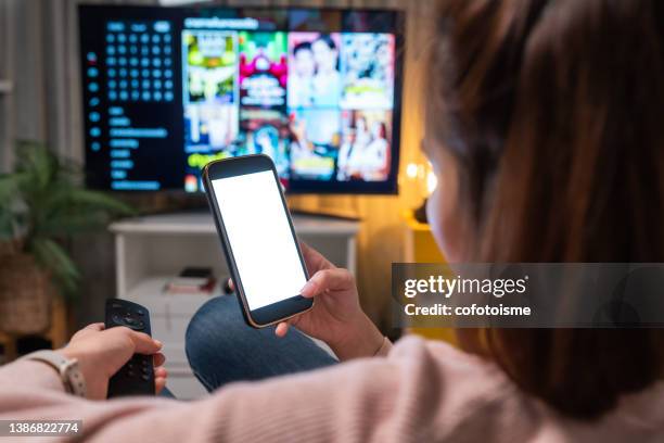 main rapprochée d’une femme tenant un téléphone avec un écran blanc vide tout en regardant la télévision à la maison - regarder tv photos et images de collection