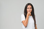 Young disappointed sad caucasian woman in white T-shirt smelling stinky, closing her nose with a hand because of odor stench isolated in grey background