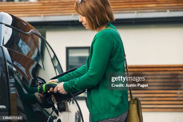 woman plugging the charging cable into her electric car as she arrives at home - elektrische auto stockfoto's en -beelden