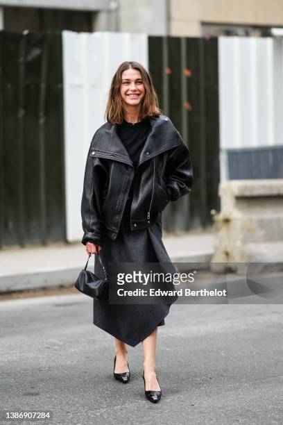 Sophia Roe wears a black pullover, a black shiny leather oversized biker jacket, a dark gray asymmetric midi skirt, a black shiny leather handbag,...