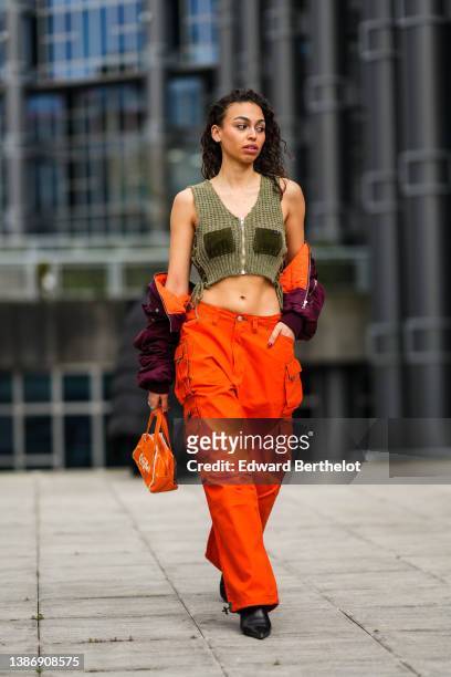 Maria Decremps wears earrings, a khaki wool braided V-neck / cropped / zipper tank-top, a purple cropped bomber coat, high waist orange cargo pants,...