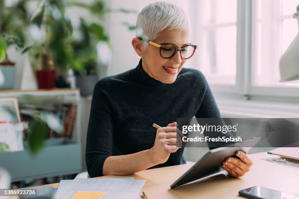 a beautiful smiling elegant senior woman reading something on her tablet while sitting at her desk in the office and working - woman reading script stock pictures, royalty-free photos & images