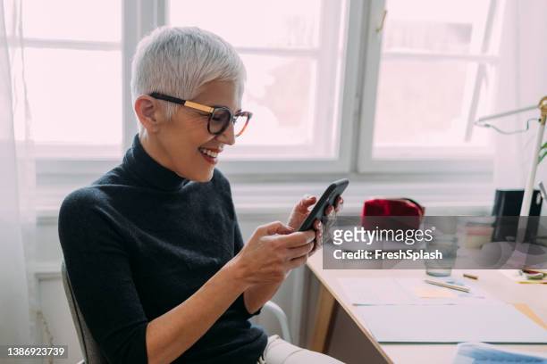 eine seitenansicht einer schönen lächelnden, eleganten seniorin, die auf ihr handy schaut, während sie an ihrem schreibtisch im büro sitzt und arbeitet - effortless stock-fotos und bilder