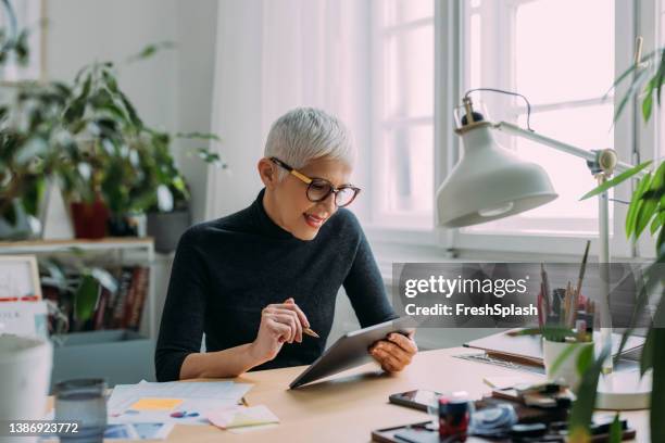 a beautiful smiling elegant senior woman looking at her tablet while sitting at her desk in the office and working - woman ipad stockfoto's en -beelden