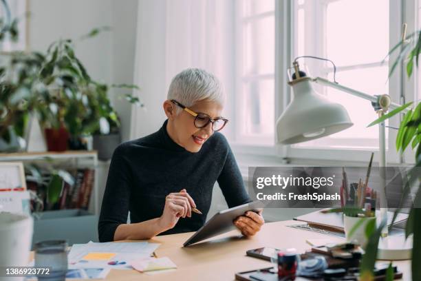a beautiful smiling elegant senior woman looking at her tablet while sitting at her desk in the office and working - woman reading script stock pictures, royalty-free photos & images