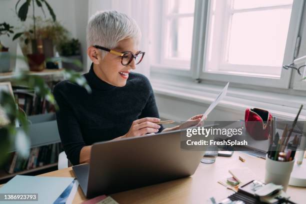 a side view of a beautiful smiling elegant senior woman looking at her notes while sitting at her desk in the office and working - journalist desk stock pictures, royalty-free photos & images