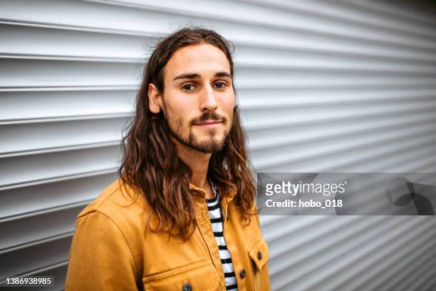 portrait d’un jeune bel homme aux cheveux longs debout dans une rue, regardant la caméra. - homme barbe fond blanc photos et images de collection