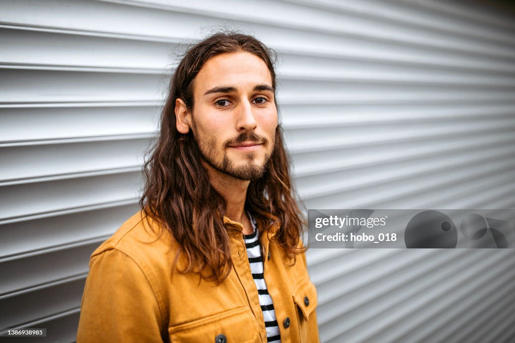 Portrait of a young handsome man with long hair standing on a street, looking at camera.