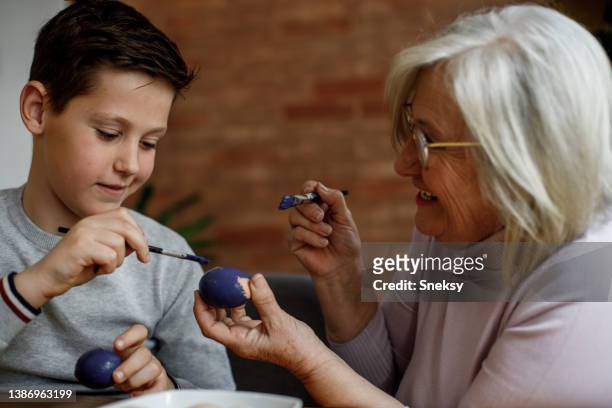 cheerful granny is using paintbrush to color easter egg in her hand. - kid boiled egg stock pictures, royalty-free photos & images