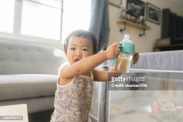 positive emotion asian baby girl holding baby bottle and drinking fresh water at home - baby cup fotografías e imágenes de stock